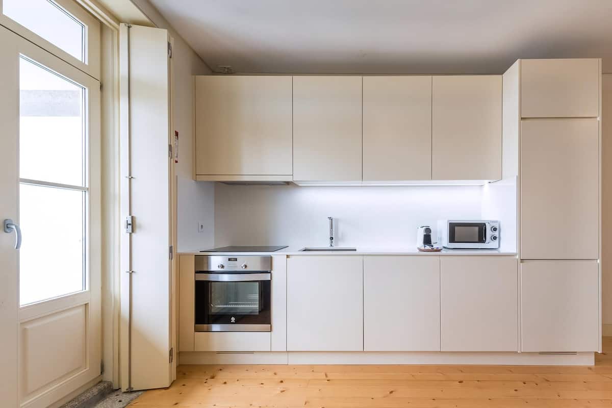 Een kleine keuken met witte kastjes, een inbouwoven, een magnetron en een raam.