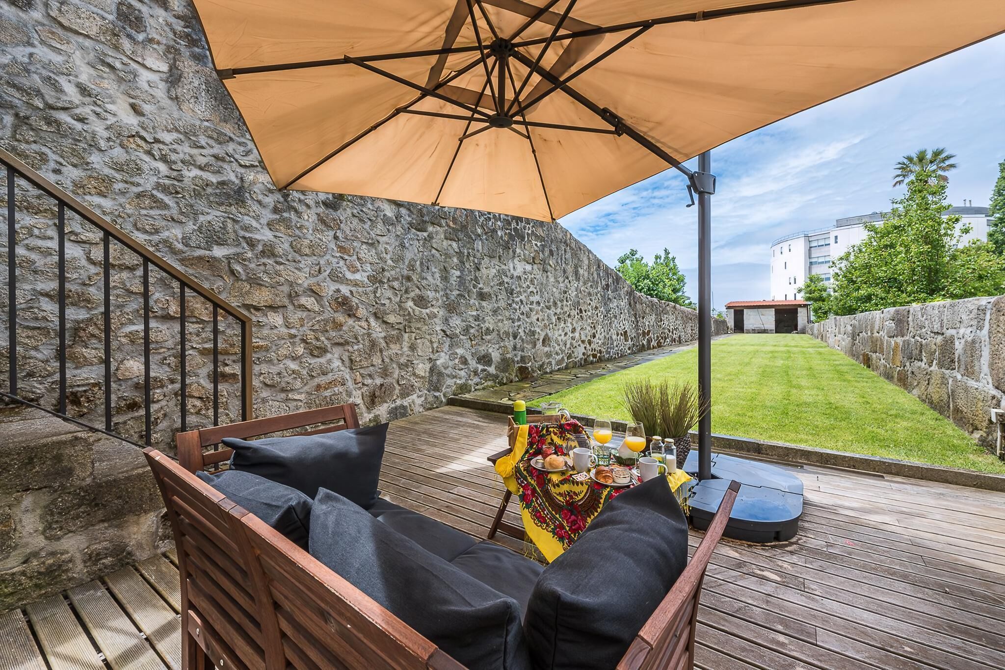 Un terrazzo in legno con un muro in pietra, un tavolo imbandito per la colazione e un ombrellone grande.