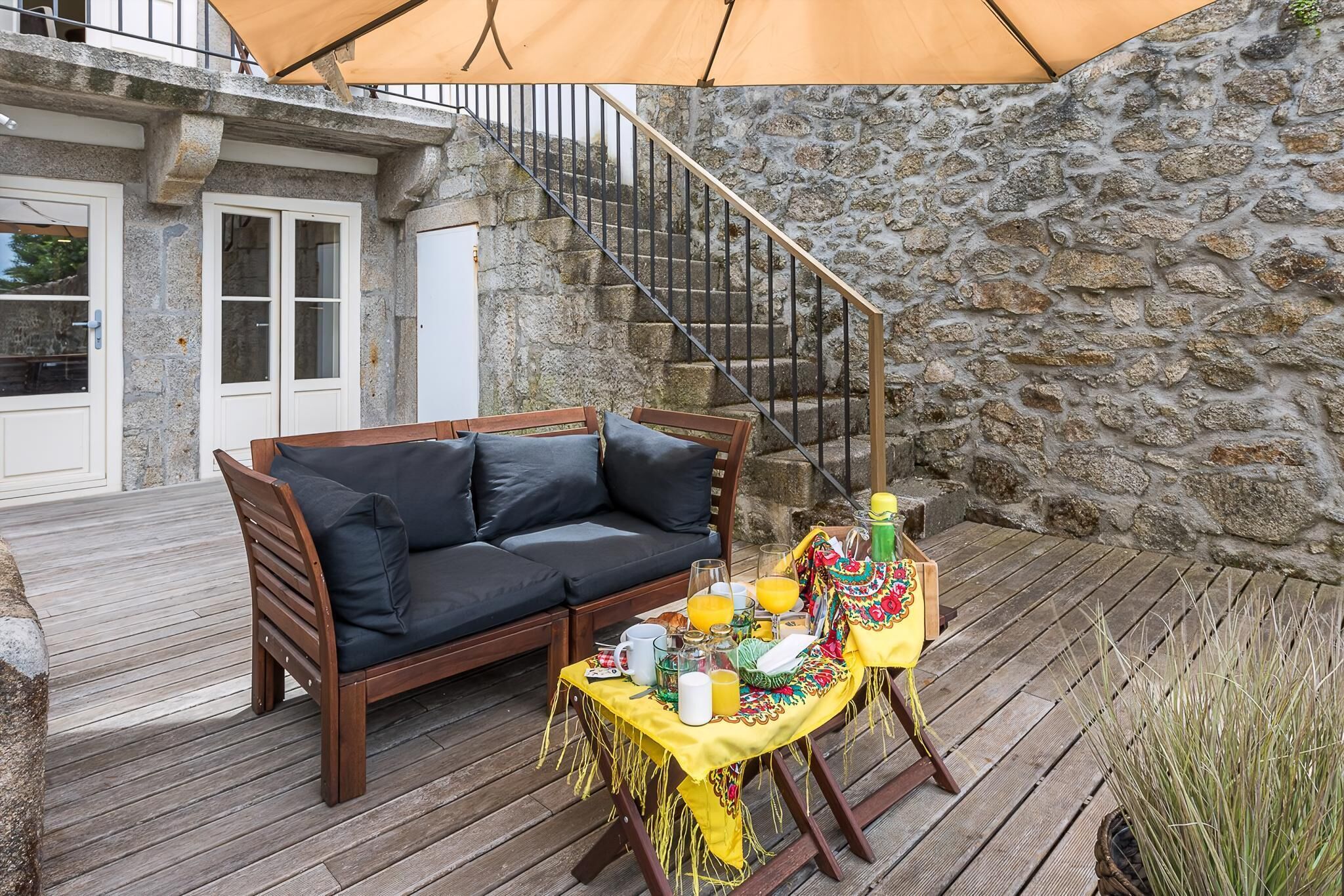 A wooden deck with a sofa and a table set for a meal, a stone wall, and a staircase.