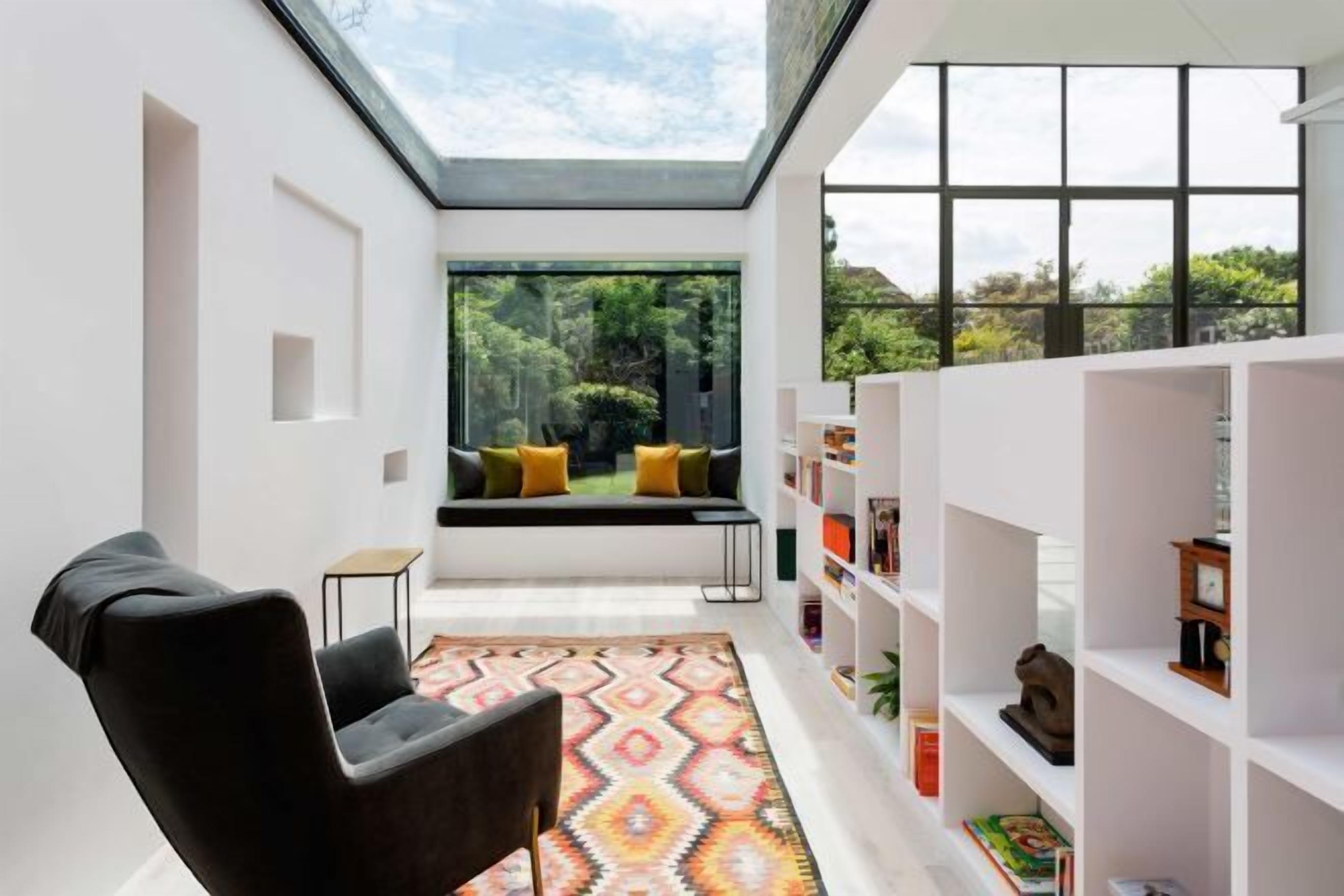 A modern interior with a skylight, a black armchair, a patterned rug, a bookshelf, and a view of greenery outside.
