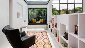 A modern interior with a skylight, a black armchair, a patterned rug, a bookshelf, and a view of greenery outside. - Quartz House (Richmond upon Thames)