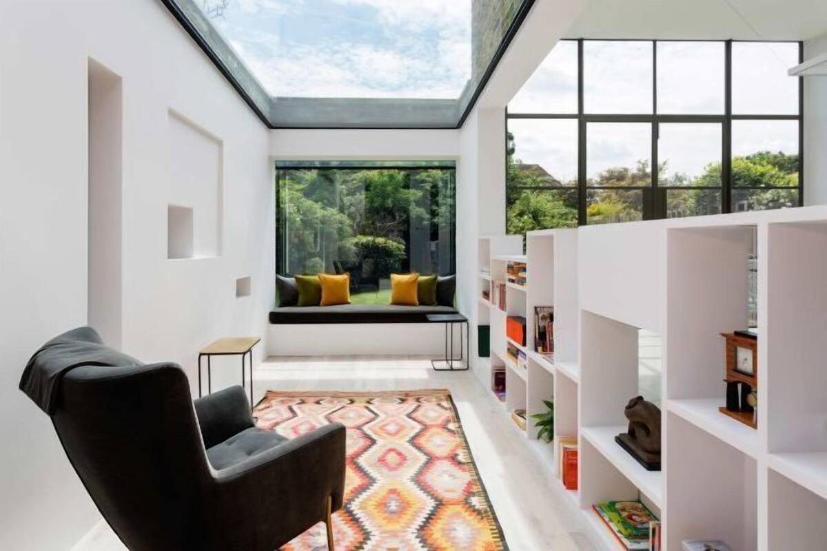A modern interior with a skylight, a black armchair, a patterned rug, a bookshelf, and a view of greenery outside.