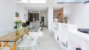 A modern kitchen with white cabinetry, a built-in oven, and a dining area with a glass table and white chairs. - Quartz House (Richmond upon Thames)