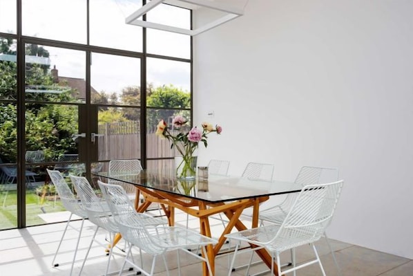 Un espace repas moderne avec une table en verre entourée de chaises blanches, un vase de fleurs et une vue sur un jardin, visible à travers de grandes fenêtres.