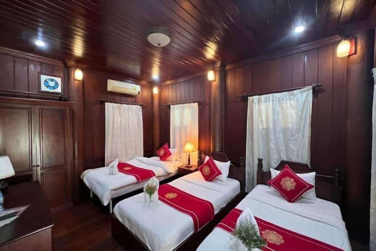 Une chambre d’hôtel avec des lambris en bois, trois lits avec une literie rouge et blanche, un bureau avec une lampe et un climatiseur fixé au mur.