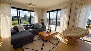 Ein modernes Wohnzimmer mit einer Couch, einem Couchtisch und einem Sessel, großen Fenstern mit Vorhängen und Blick ins Freie.