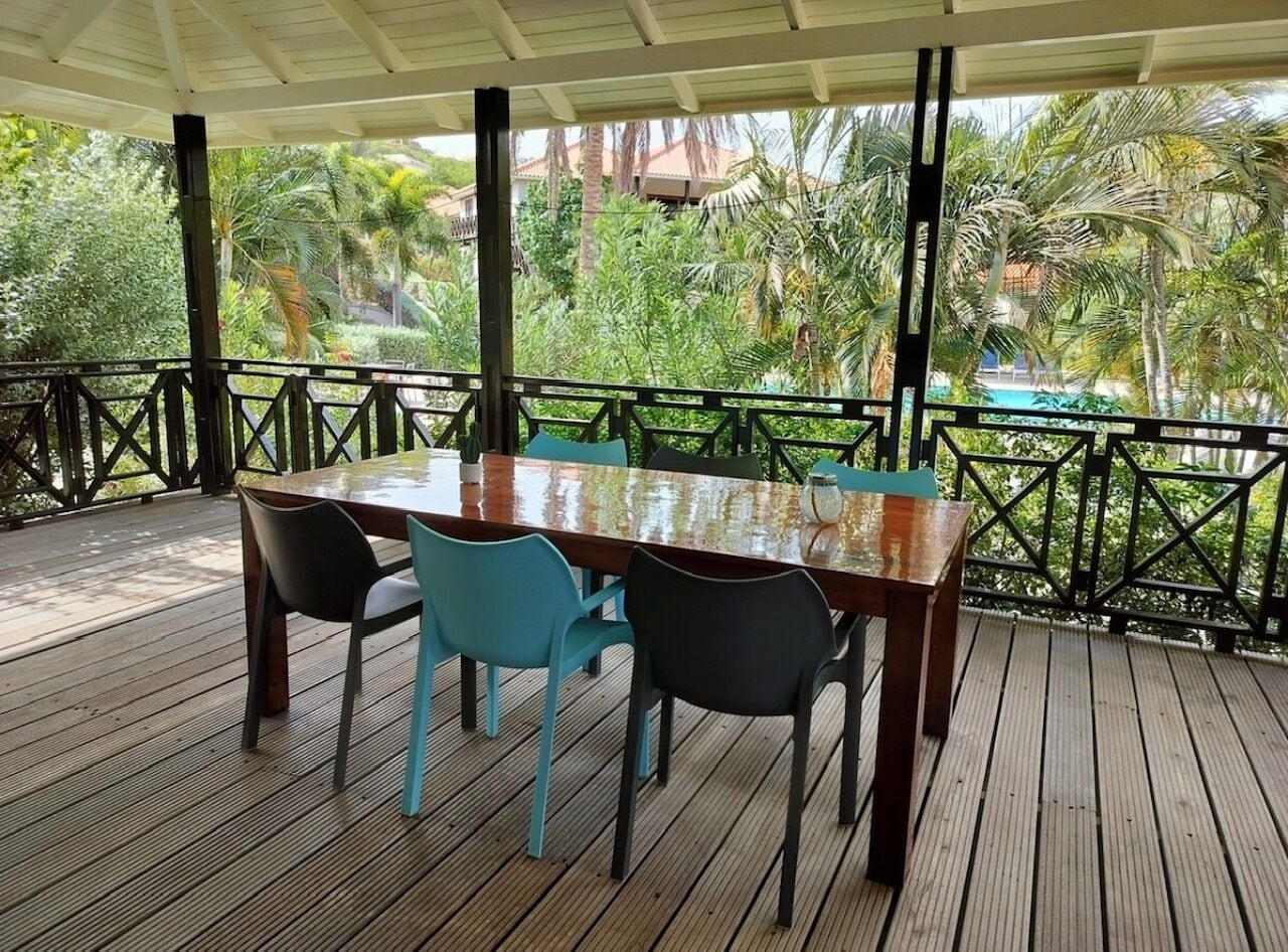 A covered outdoor dining area with a wooden table and chairs, surrounded by a railing and lush greenery.