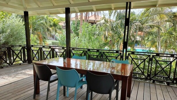 A covered outdoor dining area with a wooden table and chairs, surrounded by a railing and lush greenery. - Enjoy a spacious modern Villa, situated next to Golf Course and Pool and Beach (Sint Michiel)