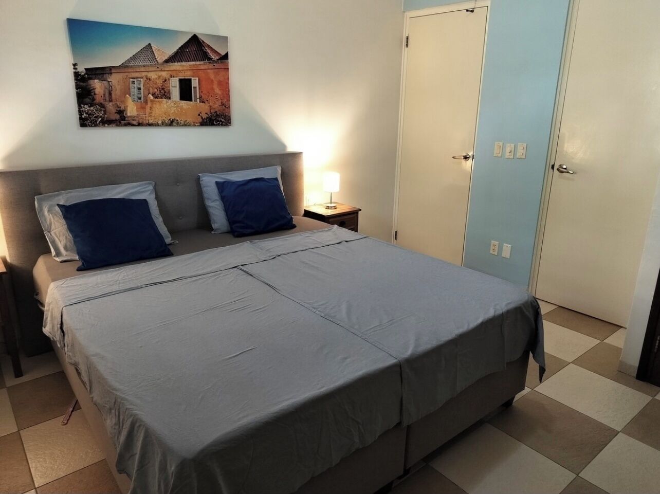 A bedroom with a bed, two nightstands, a lamp, a wall-mounted painting, and a door.