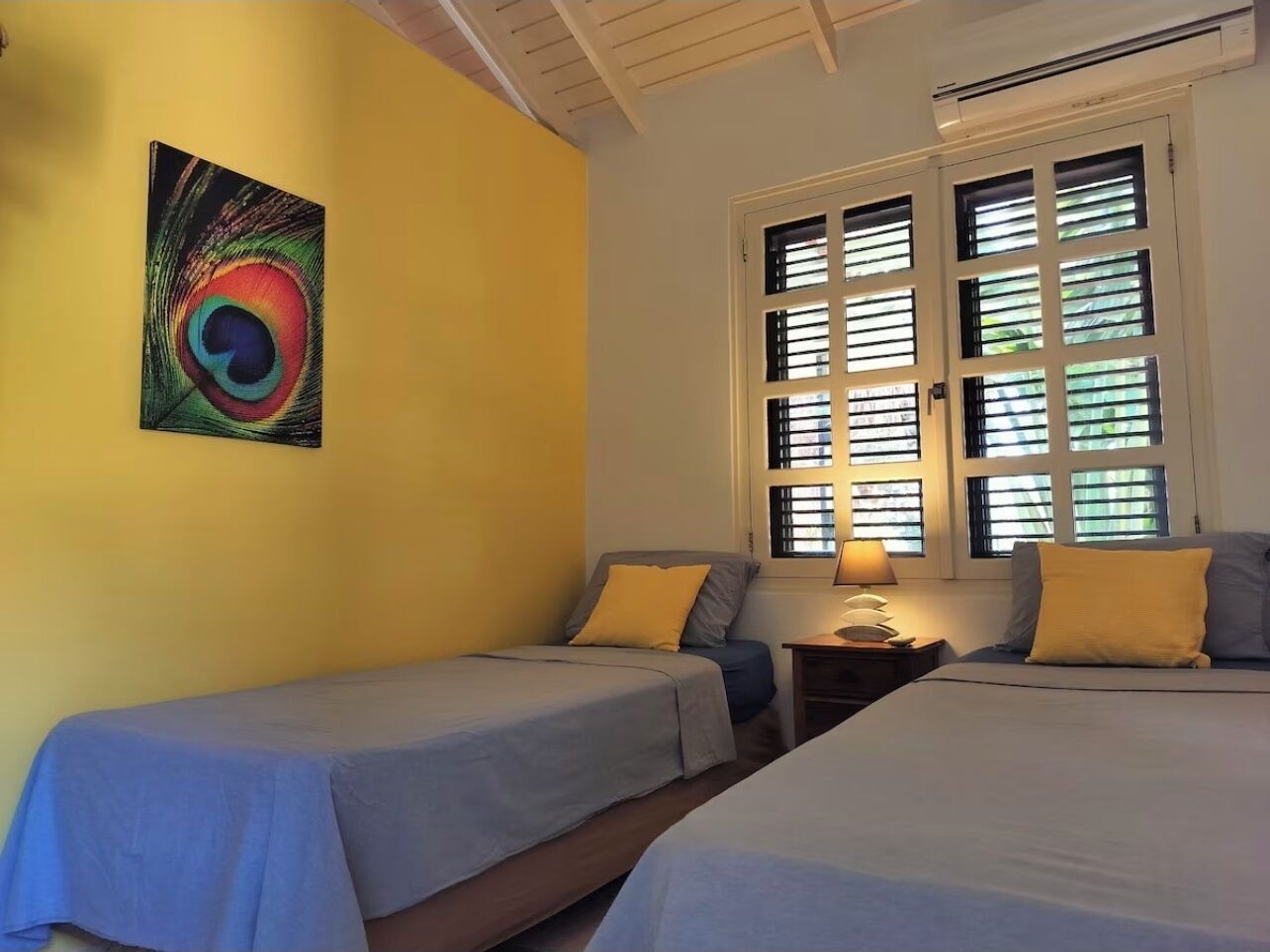 A room with two beds, a window with blinds, a wall painting, and a nightstand with a lamp.