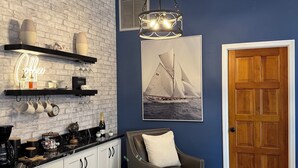 A modern kitchen with white brick walls, a wooden ceiling, and a blue accent wall. It includes a coffee bar, a dining area with a leather sofa and ottoman, and a wooden door. - Pelican’s Cove, Gulf Access, Pool, & Dock (Cape Coral)