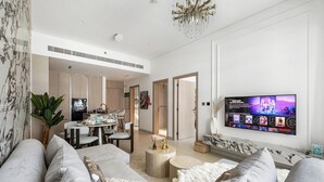 A modern living room with a large TV, comfortable seating, a dining area, and a kitchen in the background. - Stylish 1BR with PS5, Sauna and Kids` Play Area (Dubai)