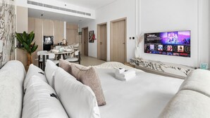 A modern living room with a sofa, a TV mounted on the wall displaying a movie, and a dining area with a table and chairs. - Stylish 1BR with PS5, Sauna and Kids` Play Area (Dubai)