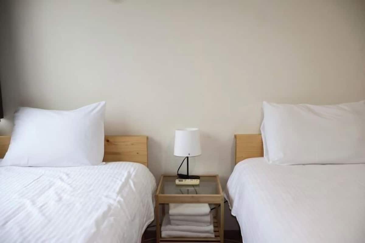 Habitació d'hotel amb dos llits individuals, cadascun amb roba de llit blanca, una petita nit de nit de fusta amb una làmpada, i una tovallola doblegada a terra.