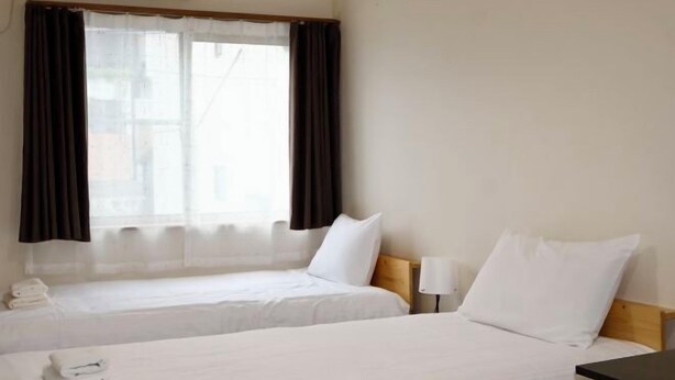 A hotel room with two beds, a window with curtains, a bedside table with a lamp, and a small table with towels.