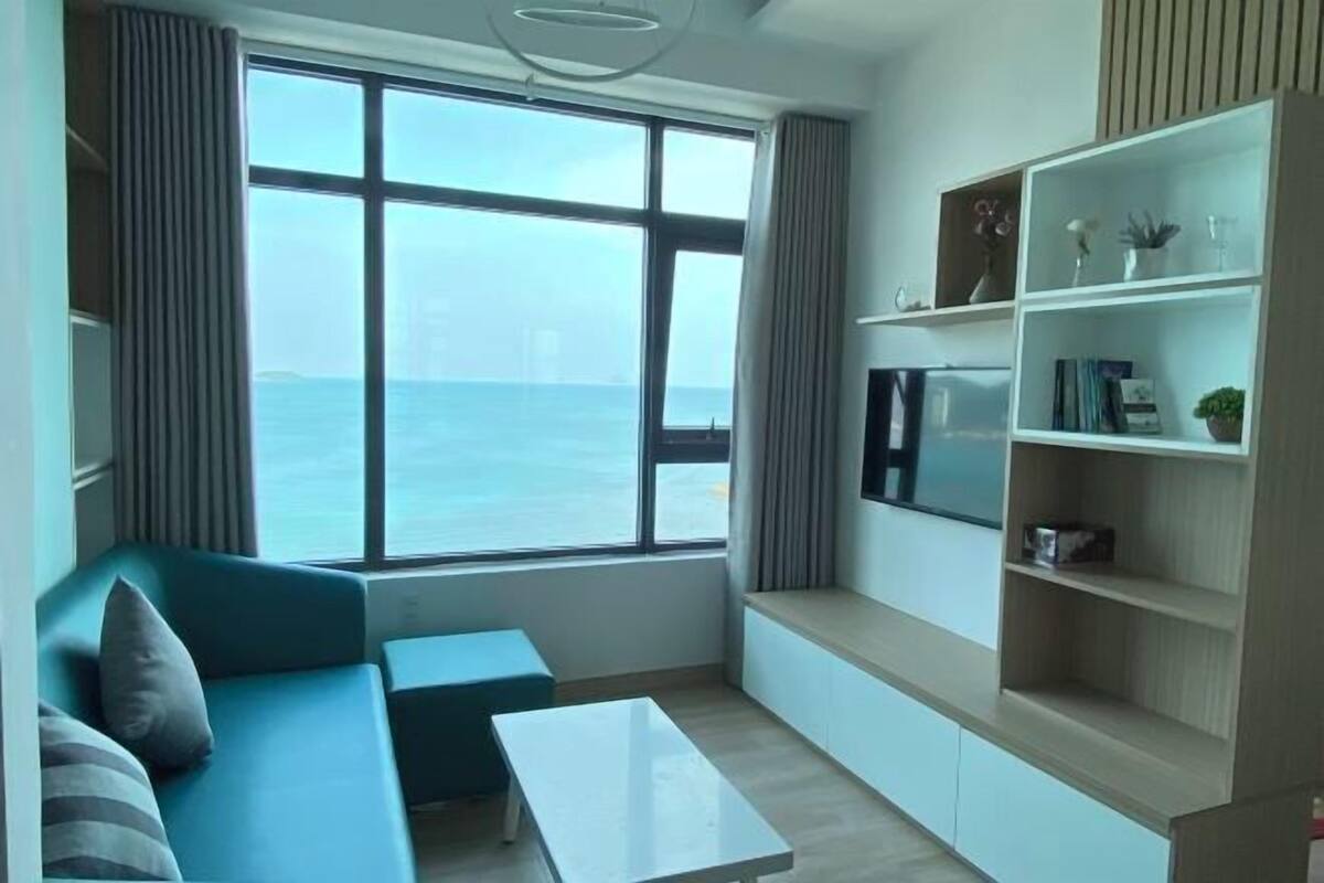 A modern living room with a large window offering a view of the sea, a blue sofa, a white coffee table, and built-in shelving with decorative items.