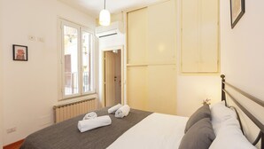 A neatly made bed with a grey blanket and white pillows, a window with blinds, an air conditioning unit, and a door leading to another room. - Rental in Rome Campo De Fiori Apartment (Rome)