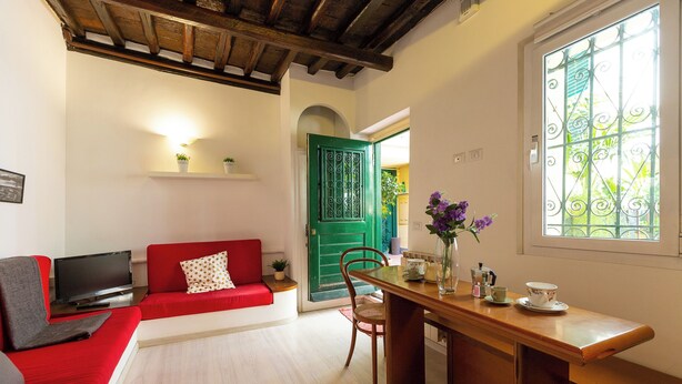 A cozy living room with a red sofa, a wooden dining table with a vase of flowers, and a green door. - Rental in Rome Campo De Fiori Apartment (Rome)