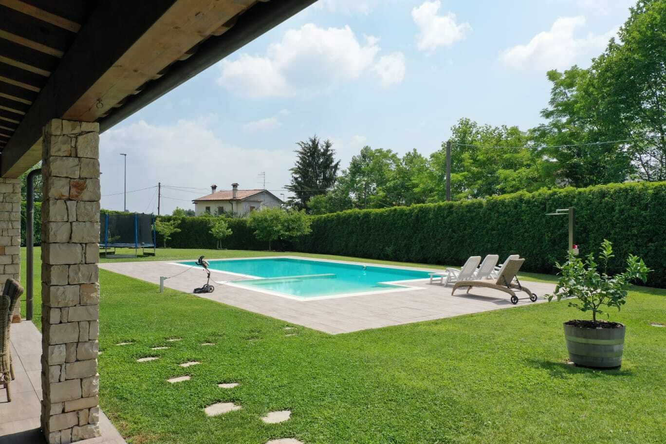 A swimming pool with a stone wall, a covered patio, and a lawn with a hedge.