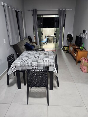 A dining area with a checkered tablecloth, black chairs, a sofa, and a television on a wooden cabinet. - Cozy MaJu Beach House (Itapoá)