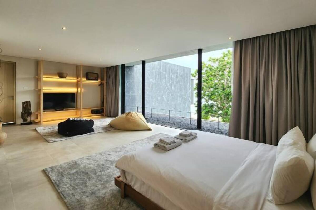 A modern bedroom with a large bed, a TV, a sofa, and a view of greenery.