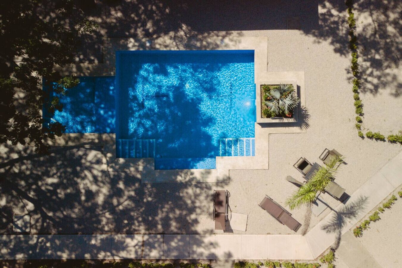 An aerial view of a rectangular swimming pool with a small deck area, surrounded by a paved walkway and shaded by trees.