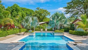 A rectangular swimming pool with a clear blue water surface, surrounded by well-maintained tropical vegetation and a small building with a sloped roof in the background. - Pool, Beach, AC, Fast WiFi | Avellanas Modern Home (Playa Avellanas)