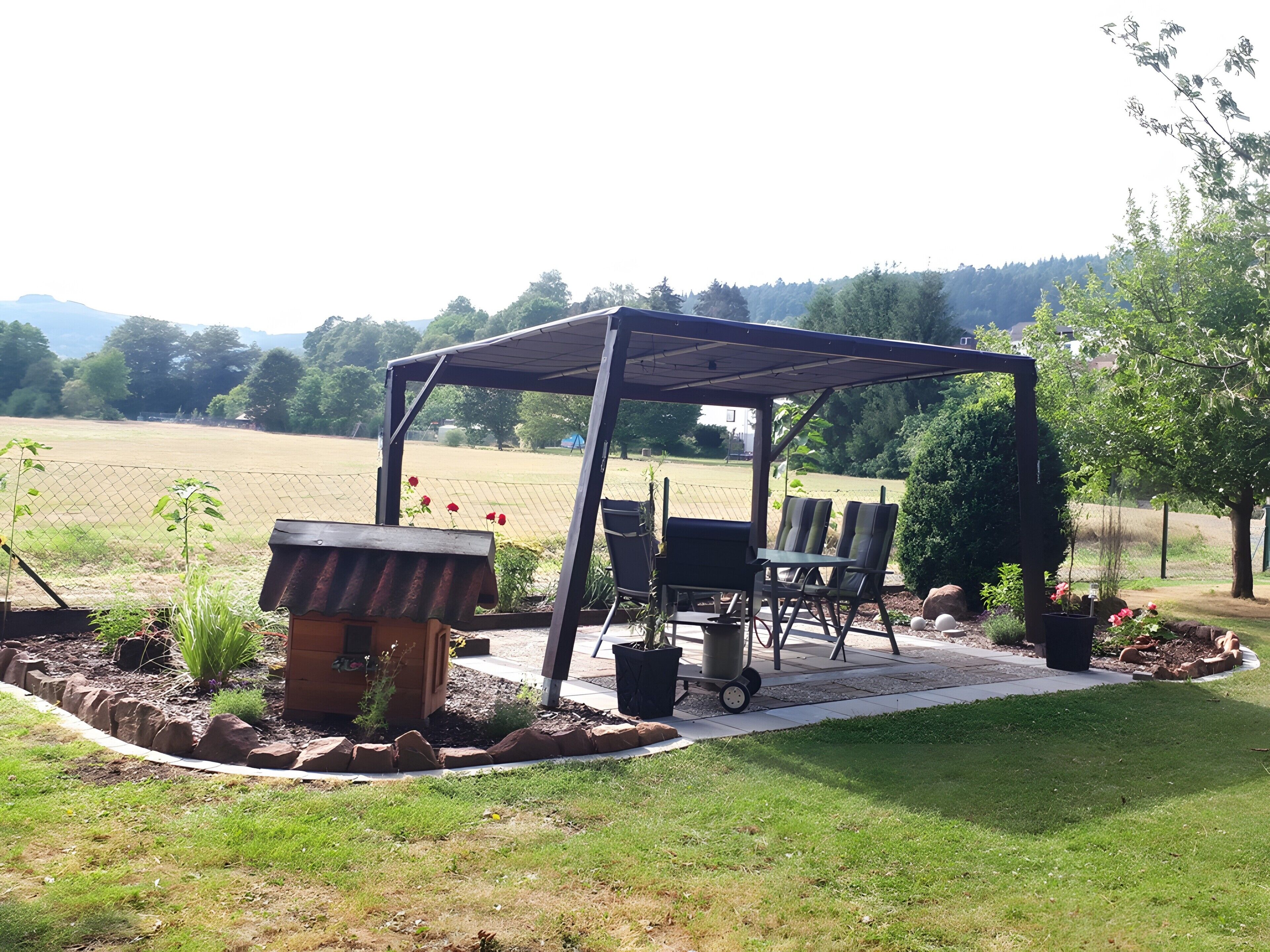 Een overdekt terras met een barbecue, stoelen en een tafel, omgeven door een tuin en uitzicht op een veld.