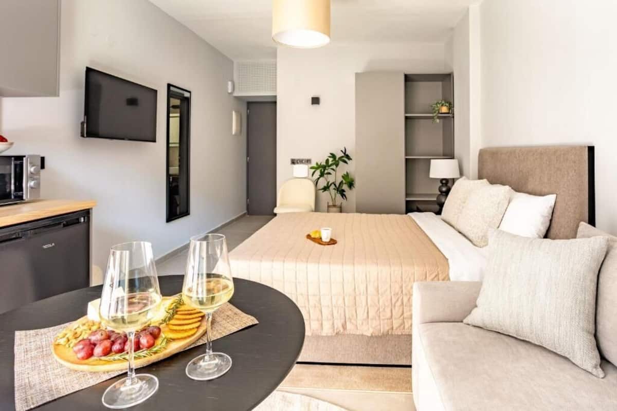 Ein Hotelzimmer mit Bett, Sofa, Wandfernseher, Schreibtisch mit Computer sowie Esstisch mit Obstteller und Gläsern.