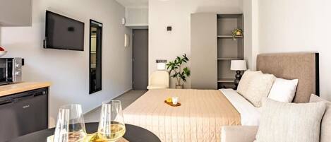 A hotel room with a bed, a sofa, a TV mounted on the wall, a desk with a computer, and a dining table with a fruit platter and glasses.