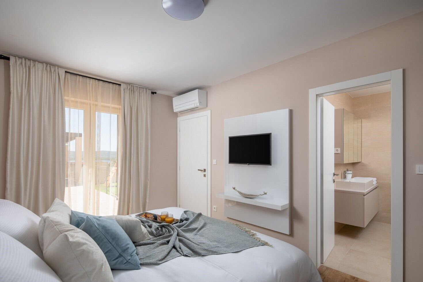 A modern hotel room with a bed, a TV mounted on the wall, a bathroom with a mirror and sink, and a balcony with curtains.