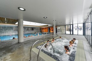 A modern indoor swimming pool area with multiple pools and a hot tub, surrounded by large windows and a high ceiling. - Résidence Privilège 4P8S (Germ)