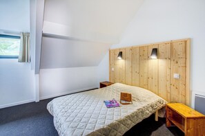 A bedroom with a wooden headboard, a quilted bedspread, a bedside table, and a window with curtains. - Résidence Privilège 4P8S (Germ)