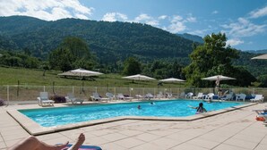 A swimming pool with lounge chairs and umbrellas, surrounded by a fence and trees, with mountains in the background.