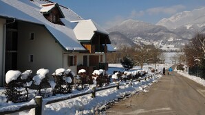 A snow-covered building with a sloped roof, a wooden porch, and a view of snow-covered mountains in the background.
