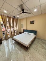 Quarto de hotel com cama, ventilador de teto, janela com cortinas, mesa de cabeceira e espelho fixado na parede.