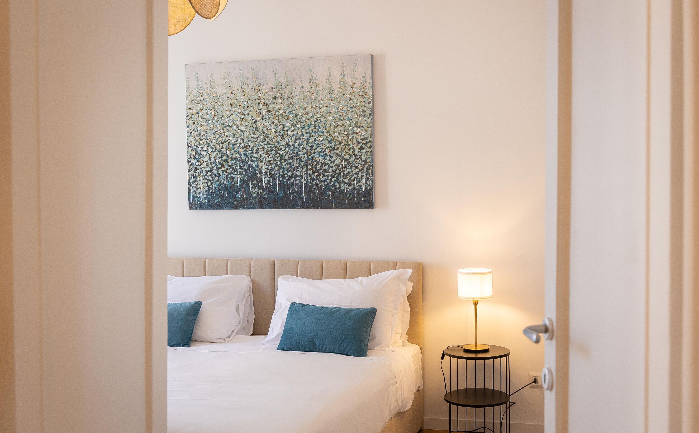 A hotel room with a bed, two pillows, a bedside table with a lamp, and a wall-mounted painting.