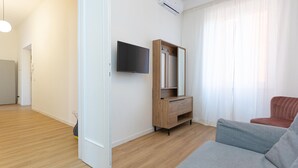 A modern living room with a wooden floor, a grey sofa, a wooden cabinet with a mirror, and a flat-screen TV mounted on the wall. - Rental in Rome Constance Apartment (Rome)