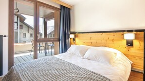 A bedroom with a wooden headboard, a large bed with white linens, a window with a view of a building, and a balcony with chairs.
