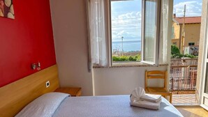 A hotel room with a bed, a chair, a window with a view of the sea, and a balcony with a railing. - Albergo Regina Mundi (Pietra Ligure)