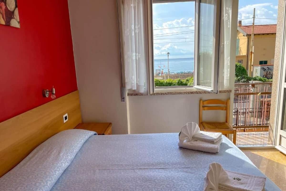 A hotel room with a bed, a chair, a window with a view of the sea, and a balcony with a railing.