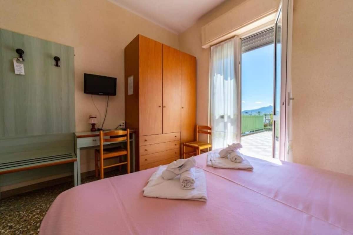 A hotel room with a bed, a wooden wardrobe, a desk with a chair, a television, and a view of the outdoors through a sliding door.