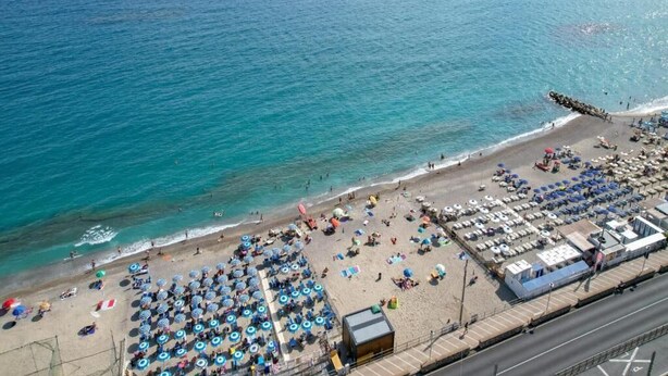 An aerial view of a beach with rows of umbrellas and sun loungers, a road parallel to the beach, and a clear view of the sea. - Albergo Regina Mundi (Pietra Ligure)