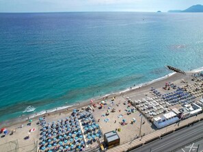 Vista aérea de uma praia com fileiras de guarda-sóis e espreguiçadeiras, uma estrada paralela à praia, e uma visão ampla do mar.