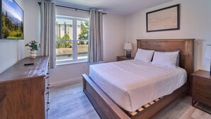 Ein Schlafzimmer mit einem hölzernen Bett, einem Schreibtisch, einem Nachttisch, einem Fenster mit Vorhängen und einem gerahmten Bild an der Wand.