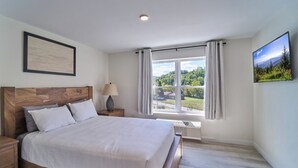 Un dormitorio con cabecera de madera, una cama grande, una mesita de noche, una lámpara, una ventana con cortinas y un cuadro enmarcado en la pared.