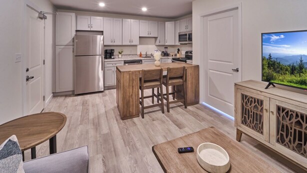 A modern kitchen with white cabinets, a stainless steel refrigerator, a wooden dining table, and a TV on a wooden cabinet. - Ruby 306: 1BD | Great LOCATION + Shared Amenities! (Pigeon Forge)