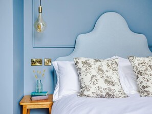 Un dormitorio con cabecera azul, ropa de cama blanca y almohadas con estampado floral.