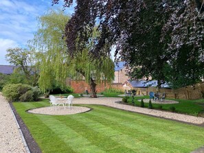 A well-maintained garden with a circular seating area, a variety of trees, and a brick wall in the background.