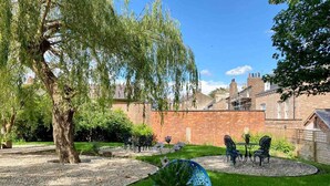 A patio with a table and chairs, surrounded by a garden with a tree and a brick wall in the background. - Railway House -Platform 5, 1 Bedroom Apartment. (York)